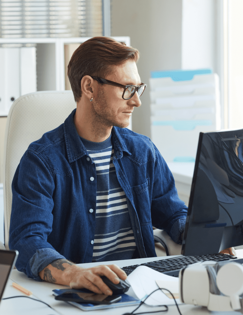 A man in glasses and casual clothing using a desktop computer at a desk.
