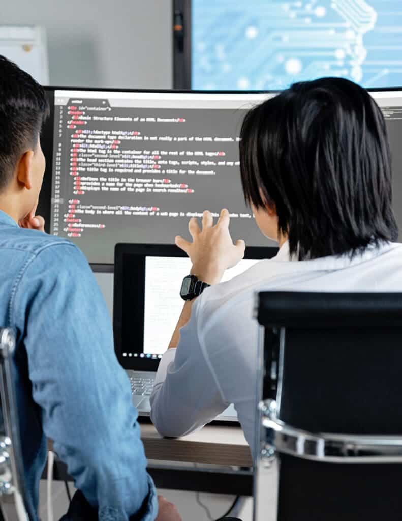 Two people look at a laptop and monitor displaying code.