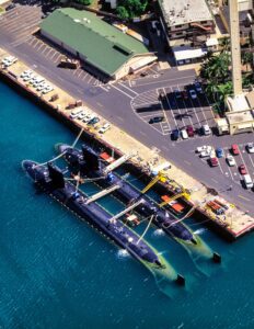 Two dark gray submarines docked on the surface of the water next to a parking lot.