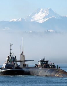 People stand on the top of a gray submarine surfaced in front of a mountain.