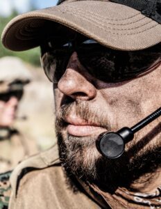 A bearded man wearing a military uniform, sunglasses, and a headset has dirt on his face.