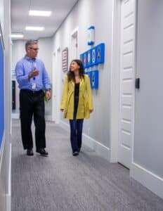 A man in a blue button-down and woman in a yellow jacket walk down the hall of an office building.