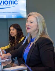 Two women in business attire sit at a glass table. One is talking and taking notes.