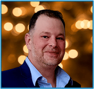 Smiling man in a suit jacket with a blurred background of warm, glowing lights.