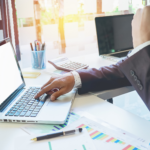 A person in a suit uses a laptop in a sunlit office. Nearby are charts, a pen, and office supplies, conveying a business and productive atmosphere.