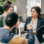 Three people engaged in conversation in an office setting, with one person gesturing and another holding documents.