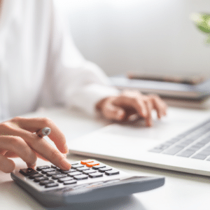 Person using a calculator and laptop at a white desk performing calculations