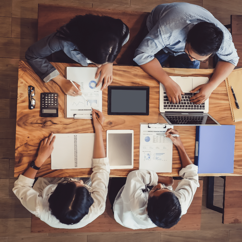 Four people working around a wooden table. Visible are notebooks, a laptop, tablet, calculator, and charts, creating a collaborative office setting.