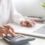 Person in a white shirt uses a calculator and a laptop on a bright desk.