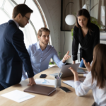 A diverse group of four professionals in a modern office gather around a laptop, engaged in discussion. The setting is collaborative and focused.