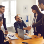 A diverse group of professionals in business attire meet in a sunlit office. A woman and a man shake hands over a table with laptops, expressing teamwork.