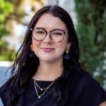 Young woman with long, dark hair and glasses, wearing a black top and gold accessories. She smiles softly, standing against a blurred outdoor backdrop.