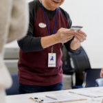 A volunteer wearing a badge and "I Voted" sticker examines an ID card at a polling station table with forms, conveying an organized, civic atmosphere.