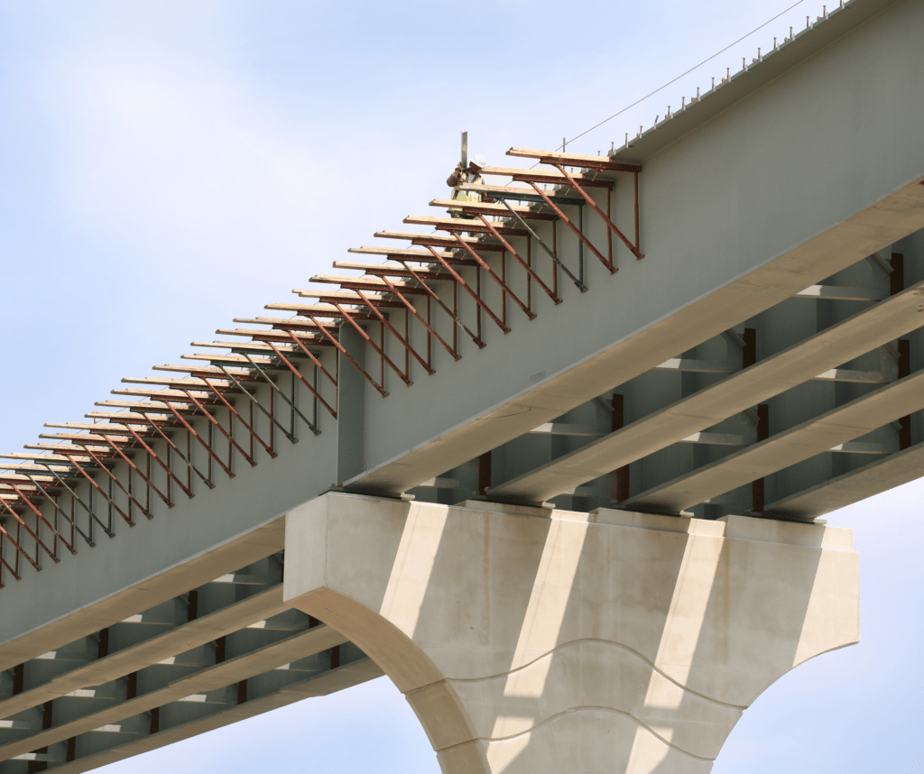 A worker in a safety vest and helmet stands on a steel beam of an unfinished bridge, surrounded by rebar supports and scaffolding against a clear sky.