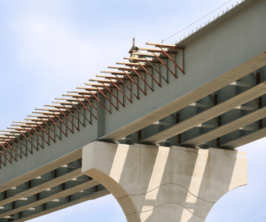 A worker in a safety vest and helmet stands on a steel beam of an unfinished bridge, surrounded by rebar supports and scaffolding against a clear sky.