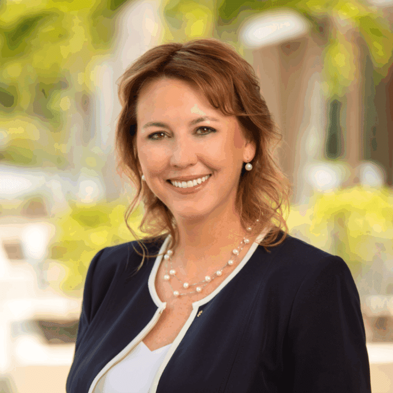 Smiling woman with light brown hair wearing a navy blazer and pearl necklace.