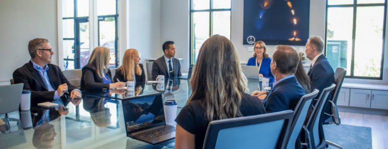A diverse group of eight professionals engaged in a meeting around a glass table in a bright, modern conference room, exuding focus and collaboration.