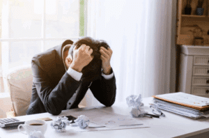 Man in a suit sits at a desk, hands gripping his hair in frustration. Crumpled papers, documents, and a coffee cup surround him, conveying stress.