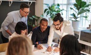A diverse group of five people collaborating around a table with documents, in a bright room filled with plants. The atmosphere is focused and cooperative.