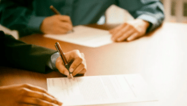 Two people seated at a wooden table sign papers, suggesting a formal agreement.