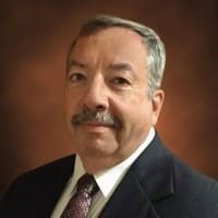 Portrait of a man with short gray hair and a mustache, wearing a black suit, white shirt, and patterned tie. He stands against a brown gradient background.