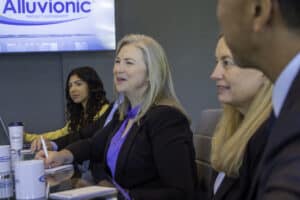 A group of professionals sitting at a conference table during a meeting at Alluvionic, with a company logo visible on the screen in the background.