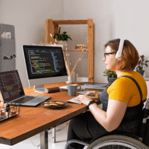 Woman in a wheelchair codes at a wooden desk with a laptop and monitor displaying colorful code.