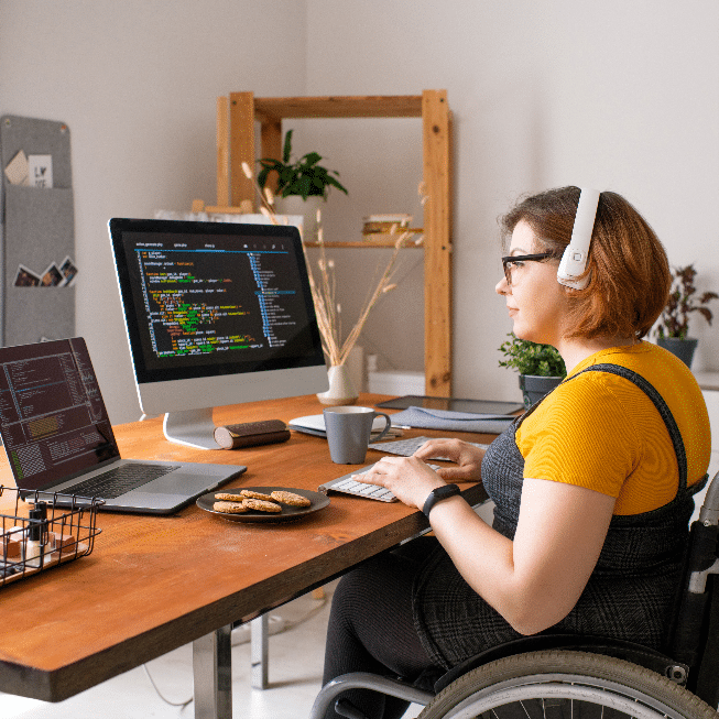 Woman in a wheelchair codes at a wooden desk with a laptop and monitor displaying colorful code.