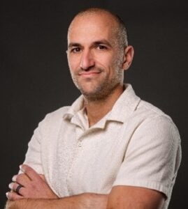 Portrait of Matt Bruggeman with arms crossed, wearing a light-colored collared shirt against a dark background.