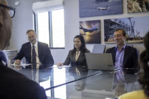 A group of business professionals sit around a glass conference table during a meeting, with one person using a laptop and others listening attentively.