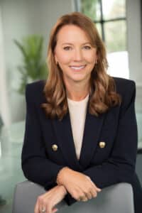 Professional headshot of a woman in a navy blazer, smiling while seated in a modern office.